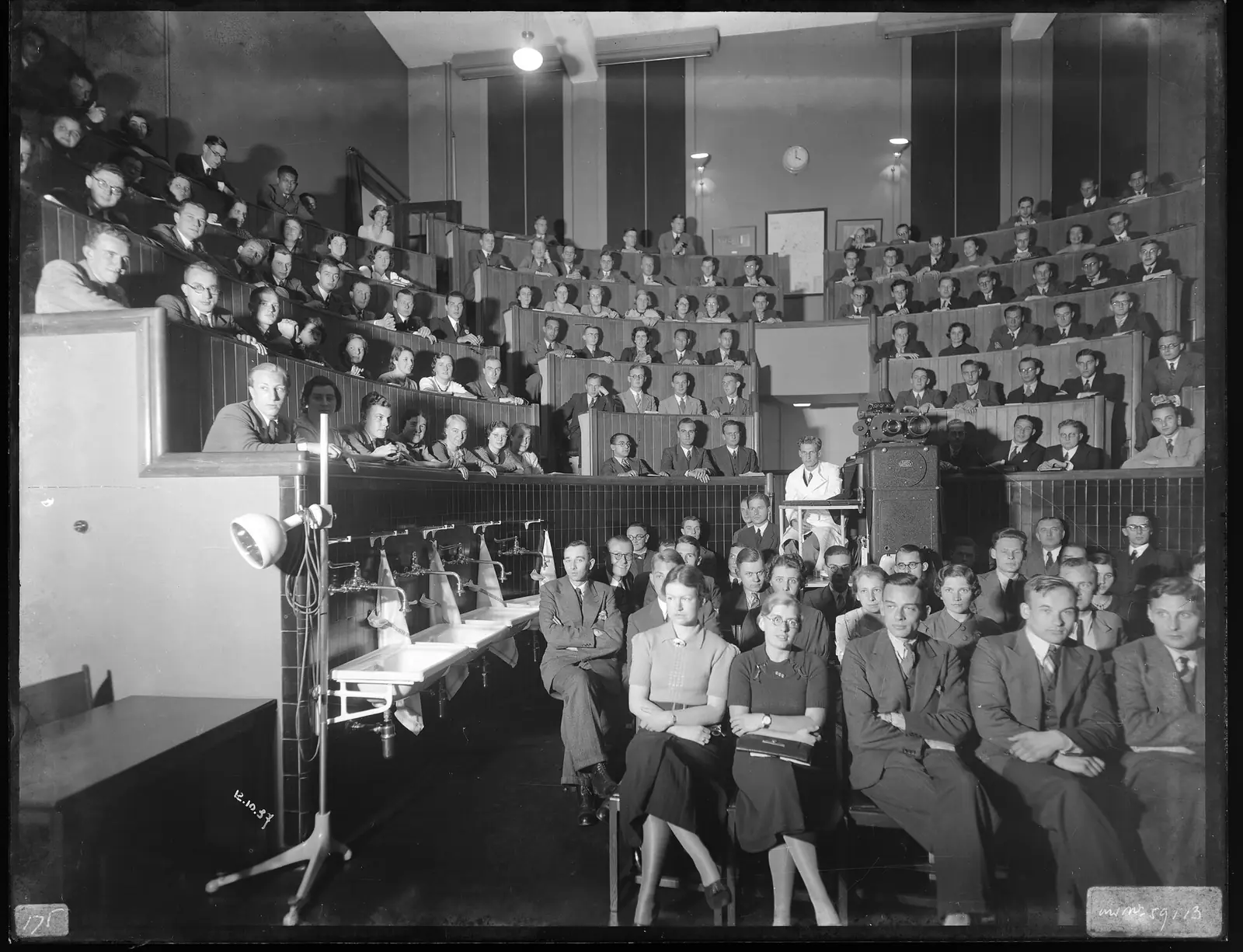 Black-and-white photograph of medical students filling a steep wooden lecture theatre at Leiden University Medical Center in 1937, with a projector and wash basins at the front